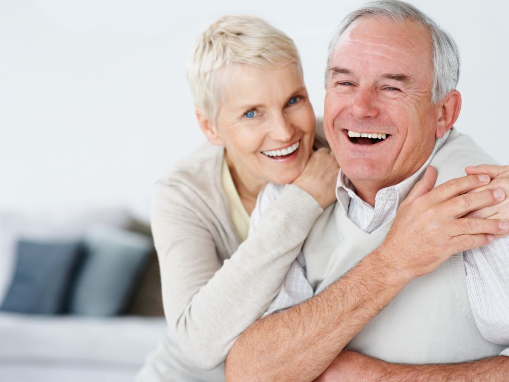 An elderly couple who is smiling with their arms around each other as their eyes face the camera.