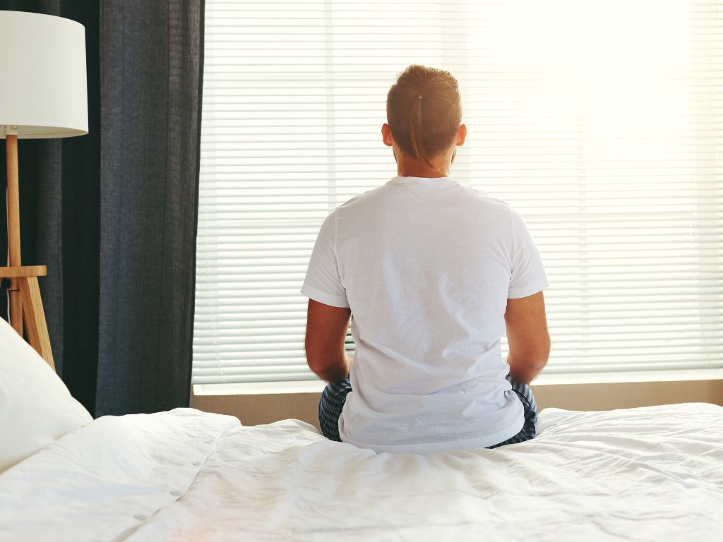 A man sitting on his bed and looking at a window with blinds down facing away from the camera